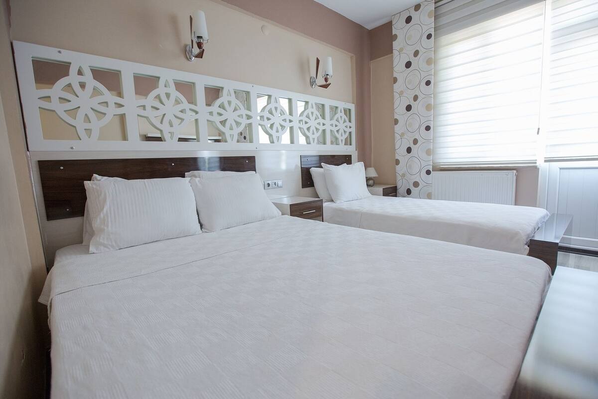 A hotel room with two beds, a decorative headboard, a nightstand, and a window with blinds.