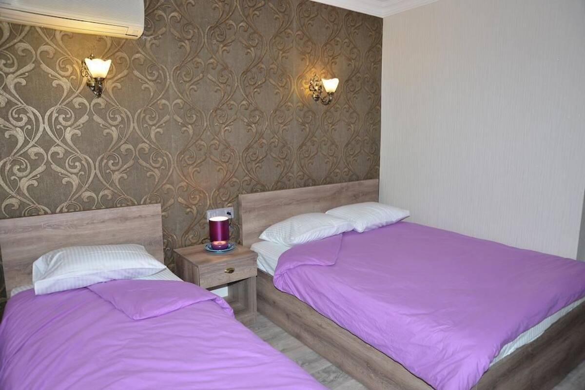 A hotel room with two beds, each with a purple bedspread and white pillows. There are two wall-mounted lamps on the patterned wallpaper.