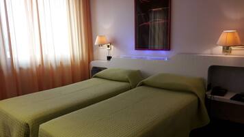 A hotel room with two single beds, a nightstand with a lamp, a wall-mounted TV, and a window with curtains.