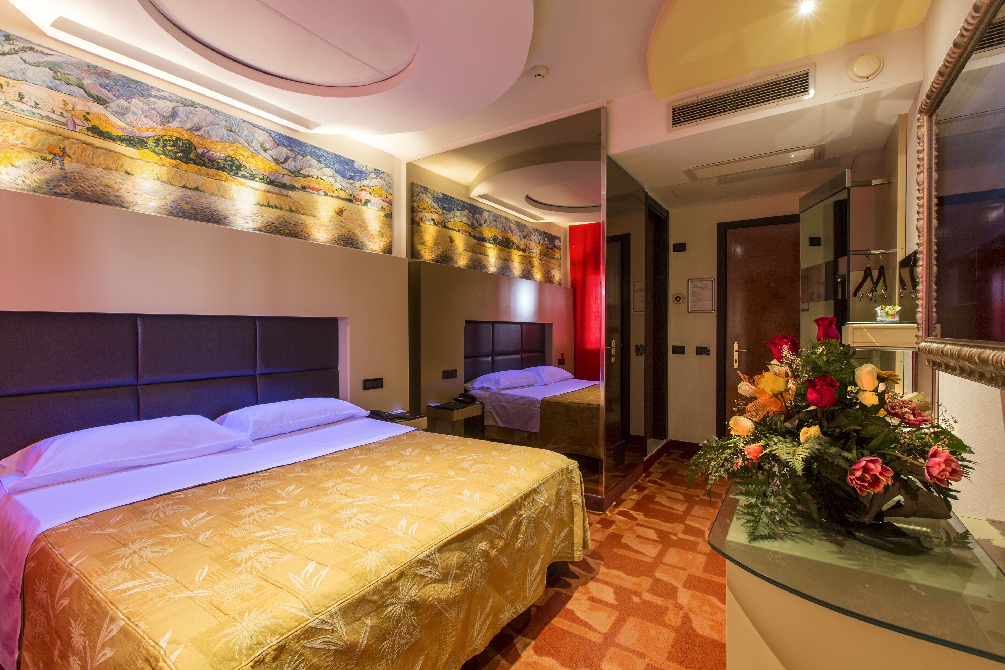 A hotel room with two beds, a large mirror, and a floral arrangement on a glass table.