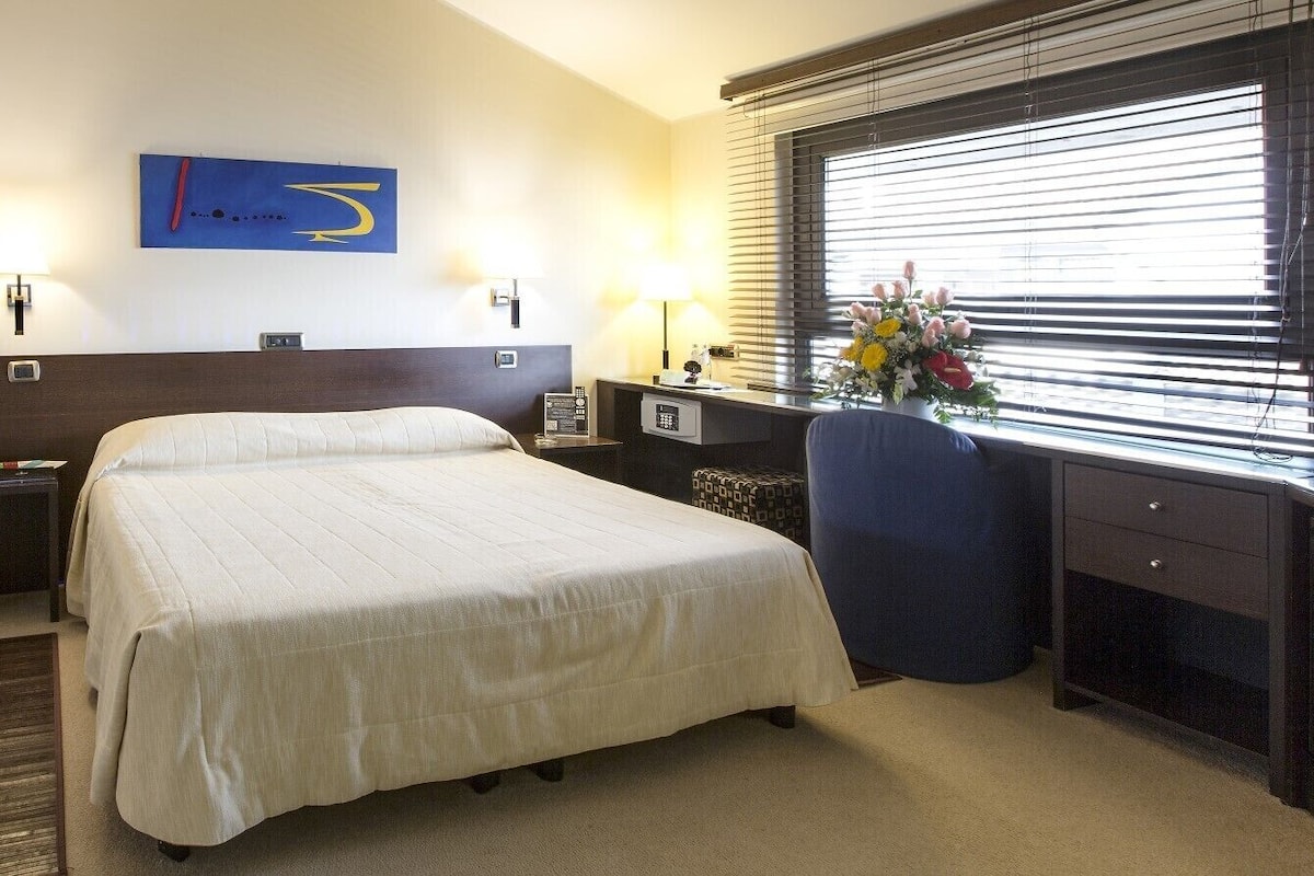 A hotel room with a large bed, bedside tables, a desk, a chair, a mirror, and a window with blinds.