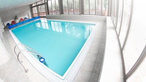 Una amplia piscina interior con agua cristalina y azul, rodeada por un suelo de baldosas y una escalera de metal.