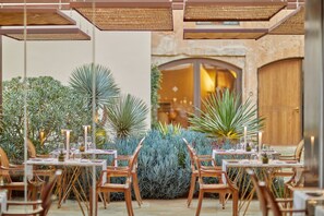 An outdoor dining area with tables and chairs, surrounded by lush greenery and a stone wall.