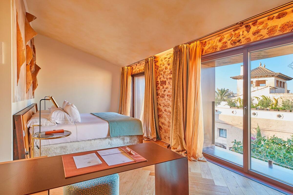 Une chambre d’hôtel avec un lit, un bureau avec des papiers, une lampe et une vue sur un immeuble à travers de grandes fenêtres.