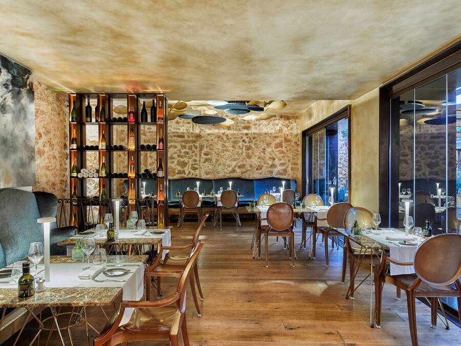 Interior de restaurante con mesas y sillas de madera, paredes de piedra y una estantería para vinos.