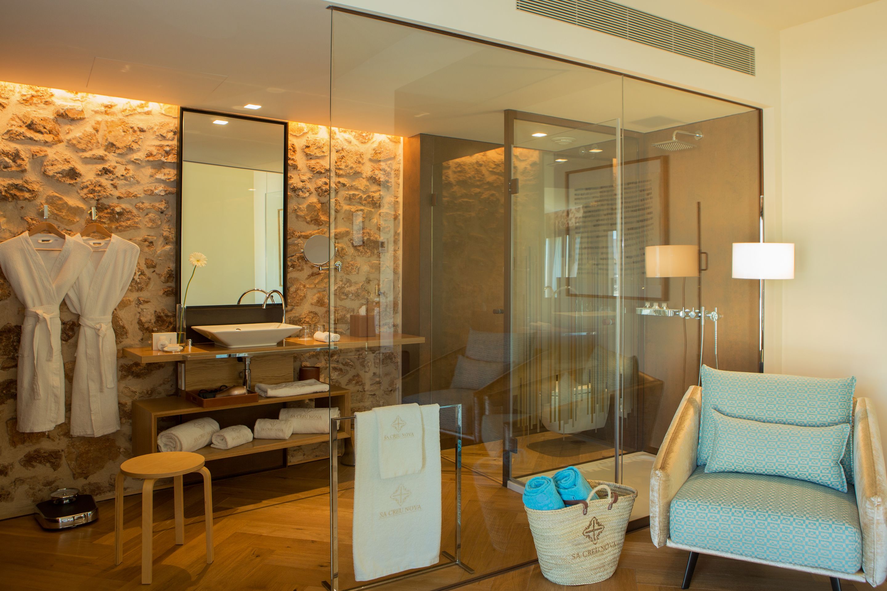 A modern bathroom with a stone wall, a glass shower enclosure, a sink, a mirror, a chair, and towels.