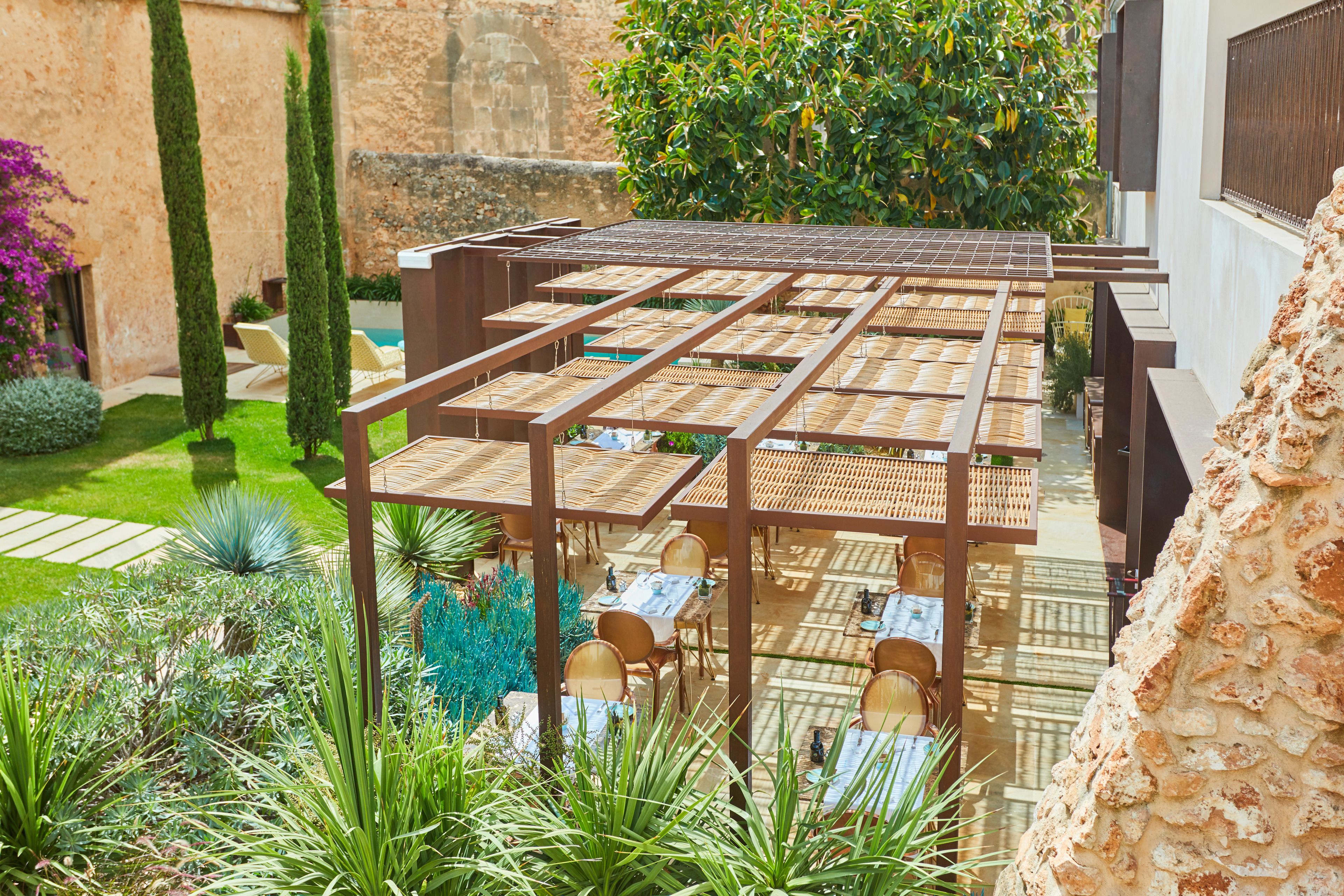 A covered outdoor seating area with tables and chairs, surrounded by greenery and a stone wall.