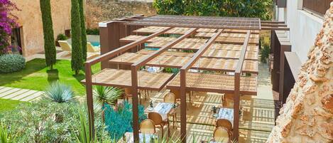 A covered outdoor seating area with tables and chairs, surrounded by greenery and a stone wall.
