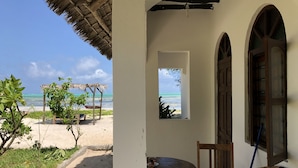 A beach view from a covered outdoor area with a table and chair, a thatched roof, and a window overlooking the ocean. - Blue Earth Beach Bungalows (Jambiani)