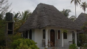 A thatched-roof building with a white exterior, surrounded by lush greenery and palm trees. - Blue Earth Beach Bungalows (Jambiani)