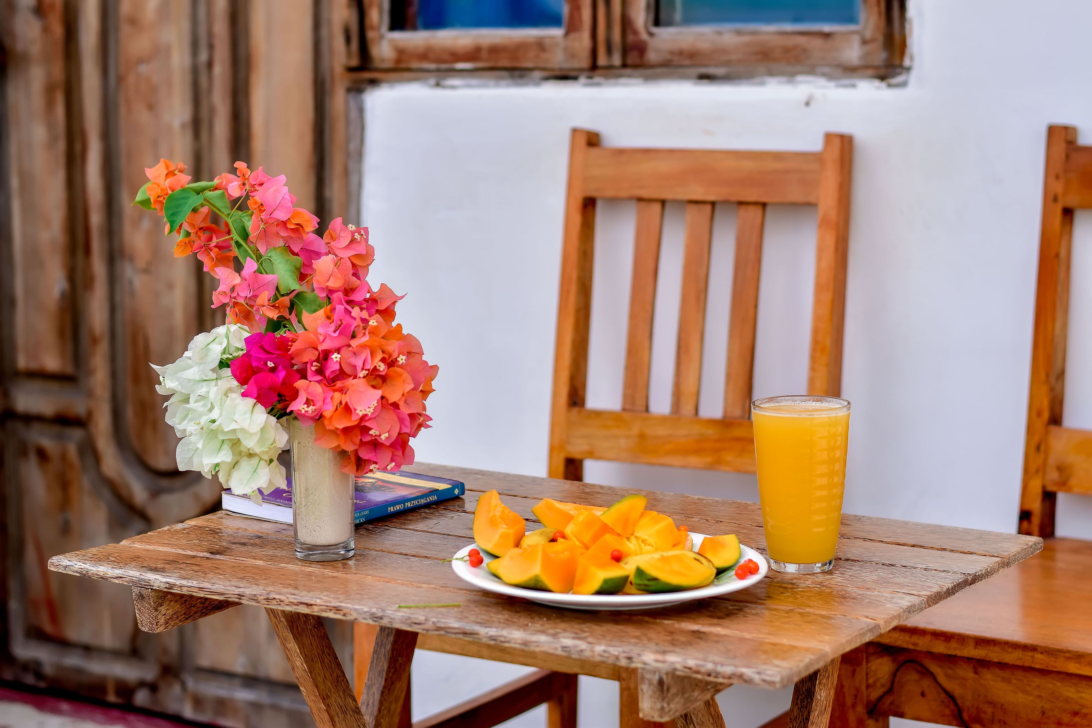 A wooden table with a plate of sliced fruit, a glass of orange juice, and a vase of flowers.