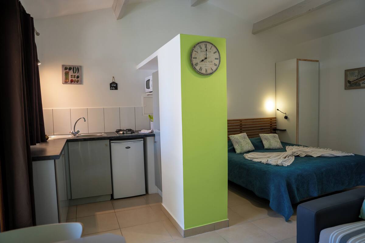 Una habitación compacta con kitchenette, una cama con cubrecama verde y un reloj de pared.