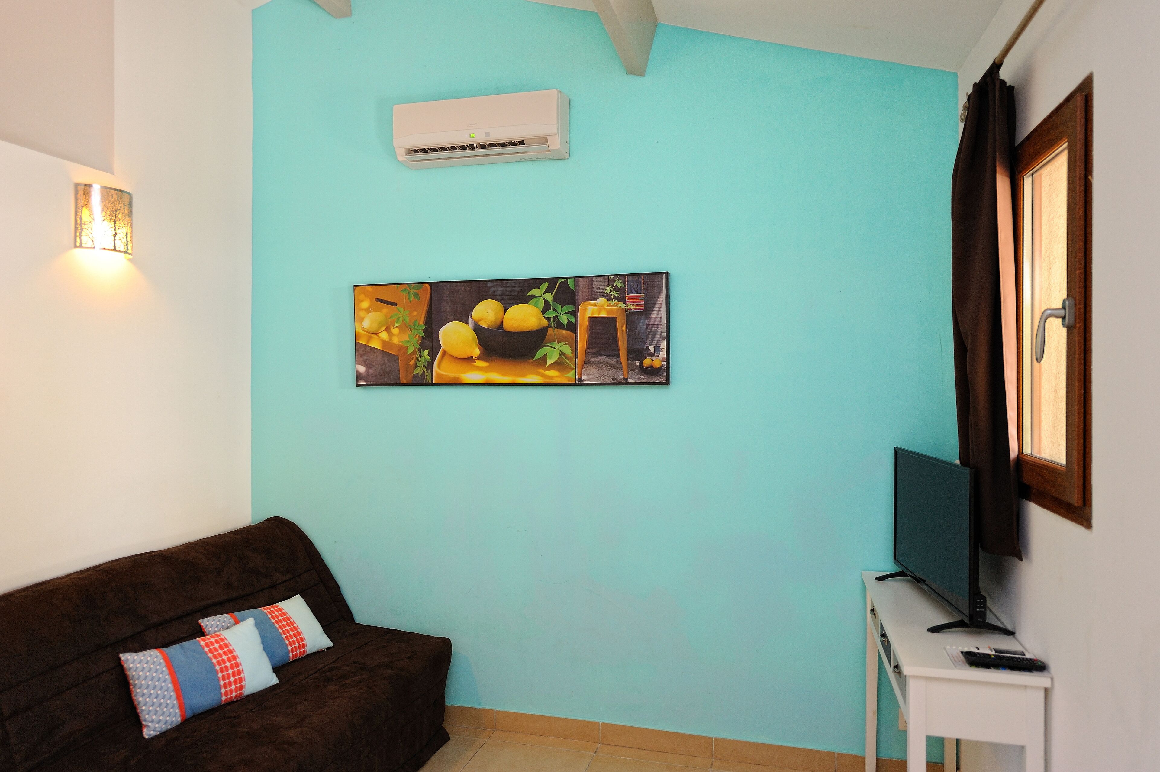 A living room with a teal wall, a brown sofa with patterned cushions, a painting of lemons, an air conditioner, a TV on a stand, and a window with curtains.