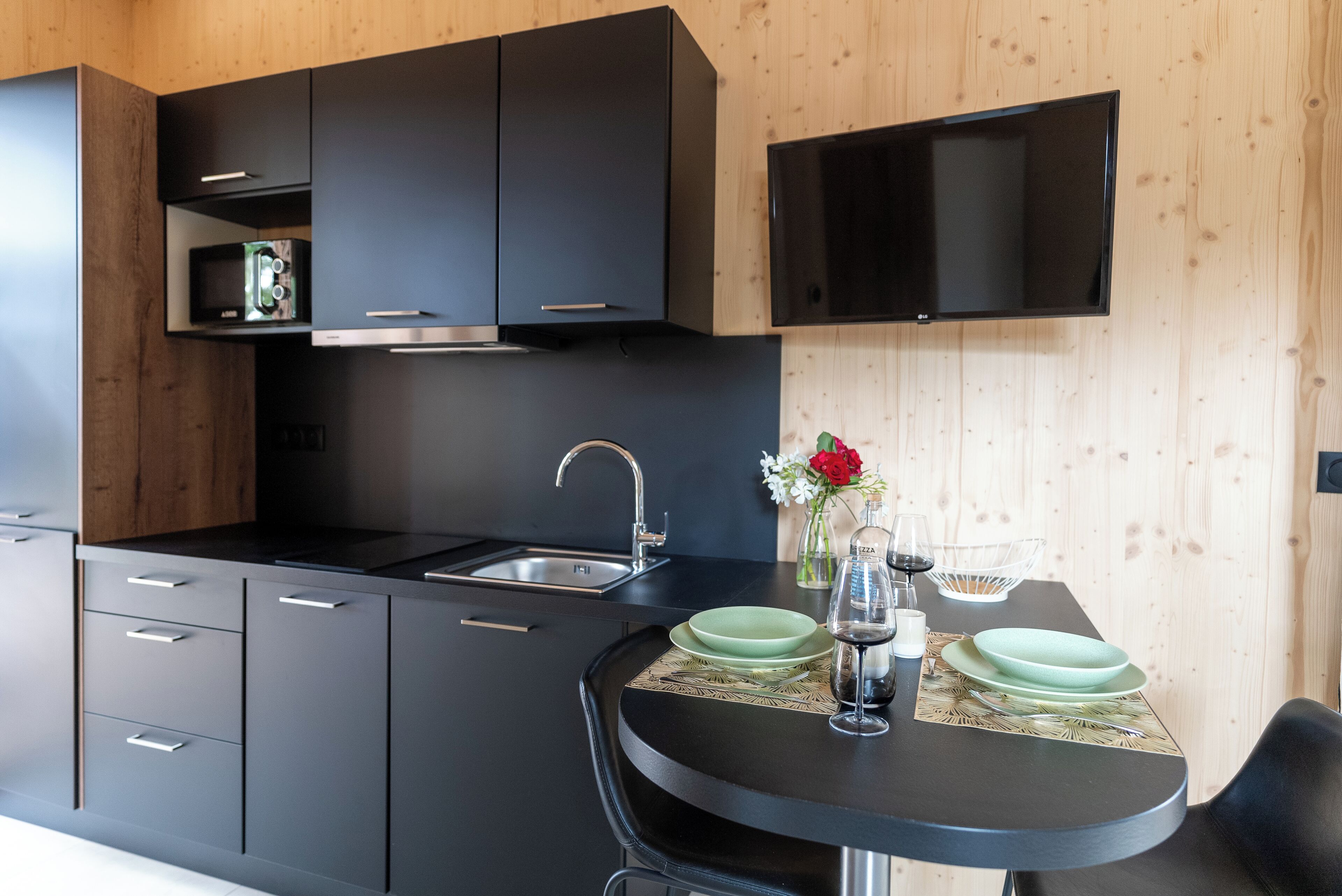 A modern kitchen with black cabinetry, a stainless steel sink, and a flat-screen TV mounted above the counter.