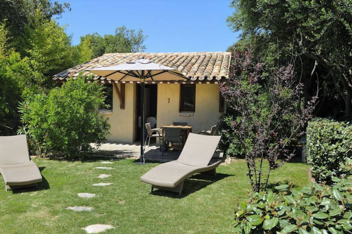 Un pequeño espacio al aire libre con dos sillones de descanso y una mesa, rodeado por un jardín con diversas plantas y una estructura de techo de tejas al fondo.