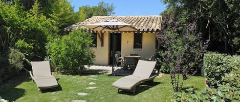 Un pequeño espacio al aire libre con dos sillones de descanso y una mesa, rodeado por un jardín con diversas plantas y una estructura de techo de tejas al fondo.