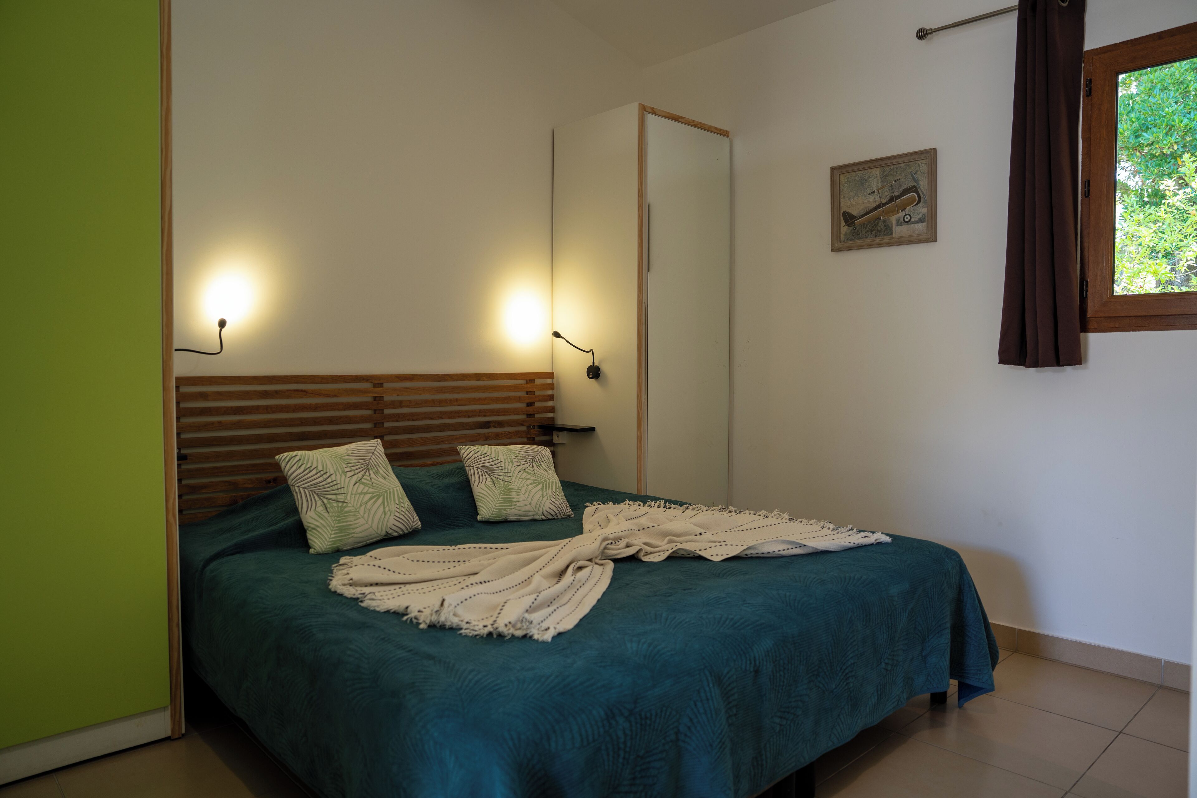 A bedroom with a bed, two wall-mounted lights, a window with curtains, a door, and a framed picture on the wall.