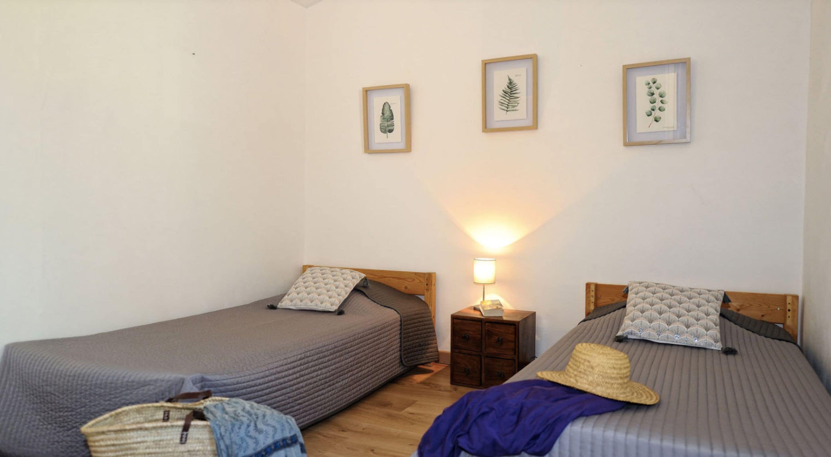 A bedroom with two single beds, a bedside table with a lamp, and three framed botanical prints on the wall.