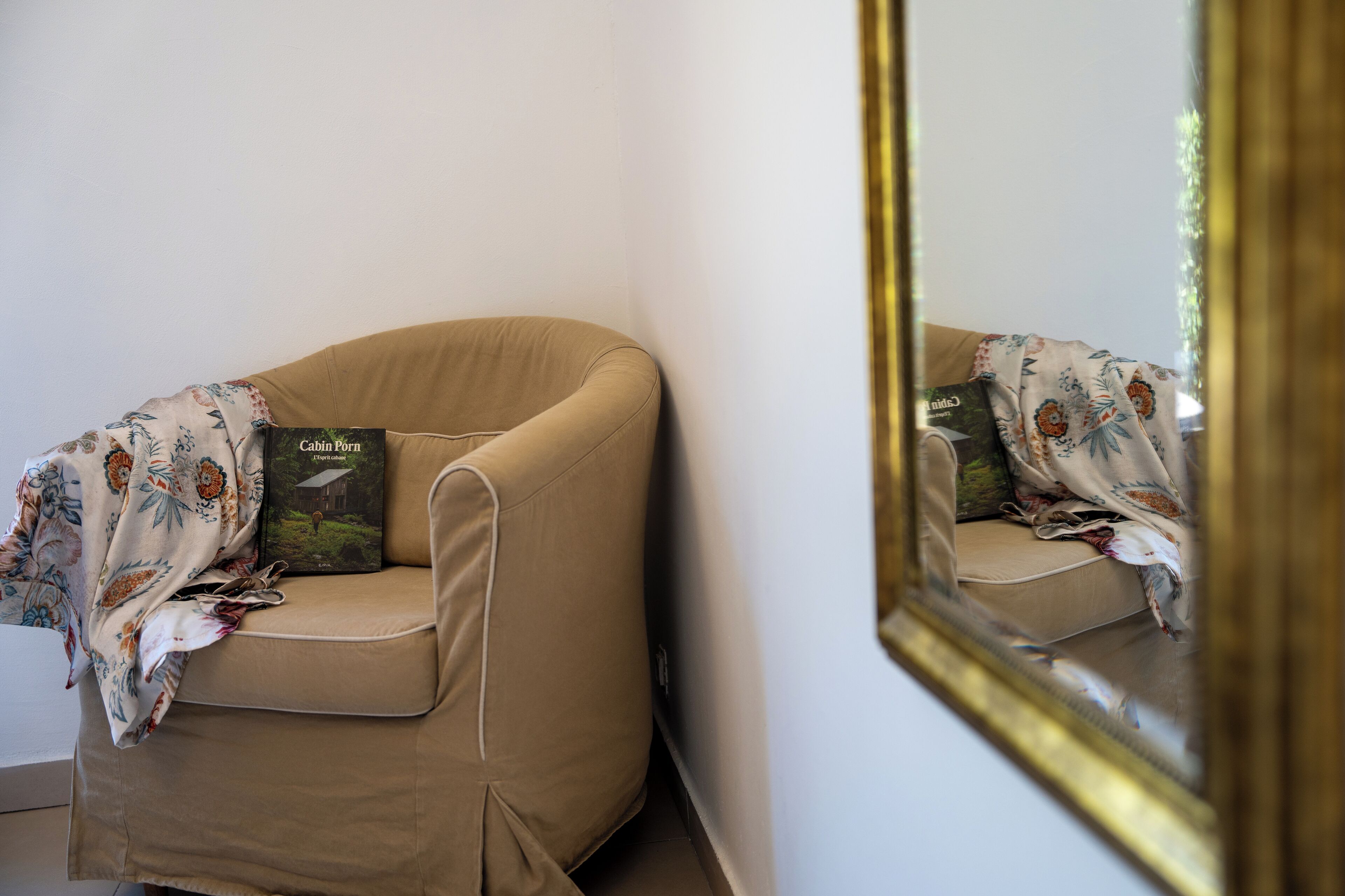 A corner of a room with a beige armchair, a floral throw blanket, and a book titled "Cabin Porn."