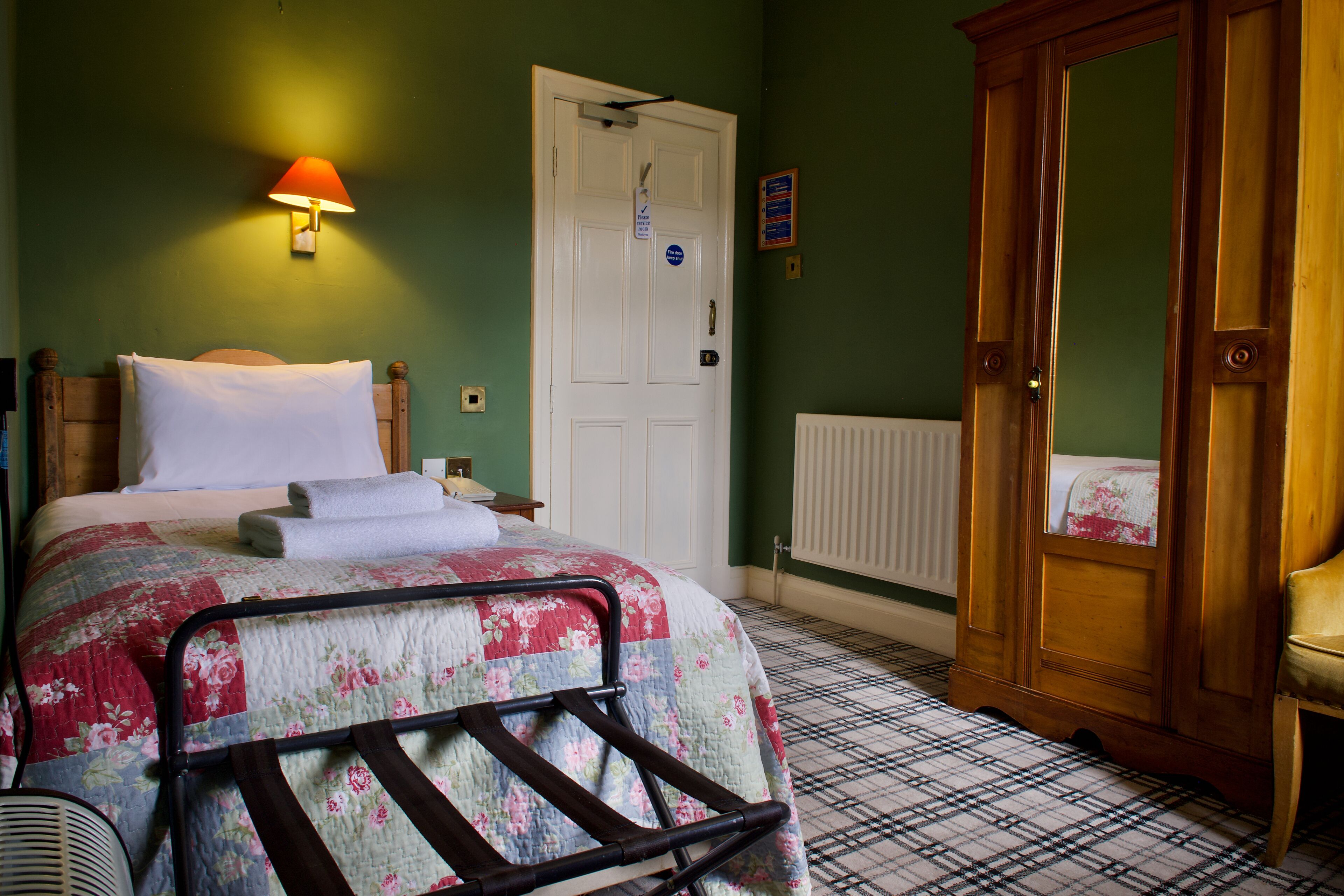 A bedroom with a bed, a nightstand, a wooden wardrobe, a radiator, and a wall-mounted lamp.