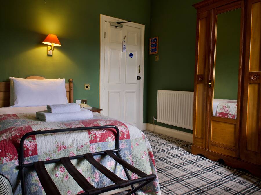 A bedroom with a bed, a nightstand, a wooden wardrobe, a radiator, and a wall-mounted lamp.
