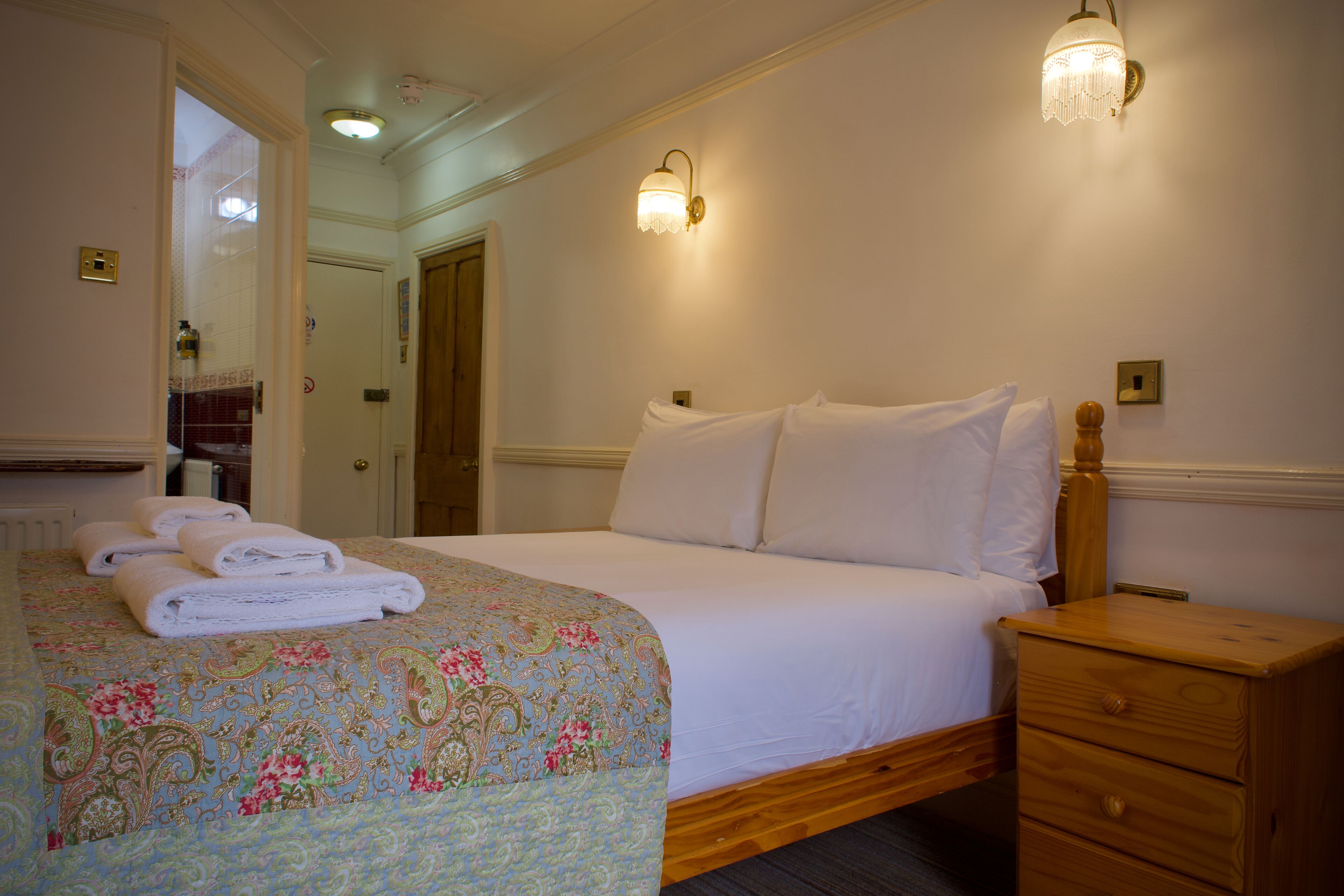 A hotel room with a bed, a nightstand, two wall sconces, a wooden nightstand drawer, and a door leading to another room.