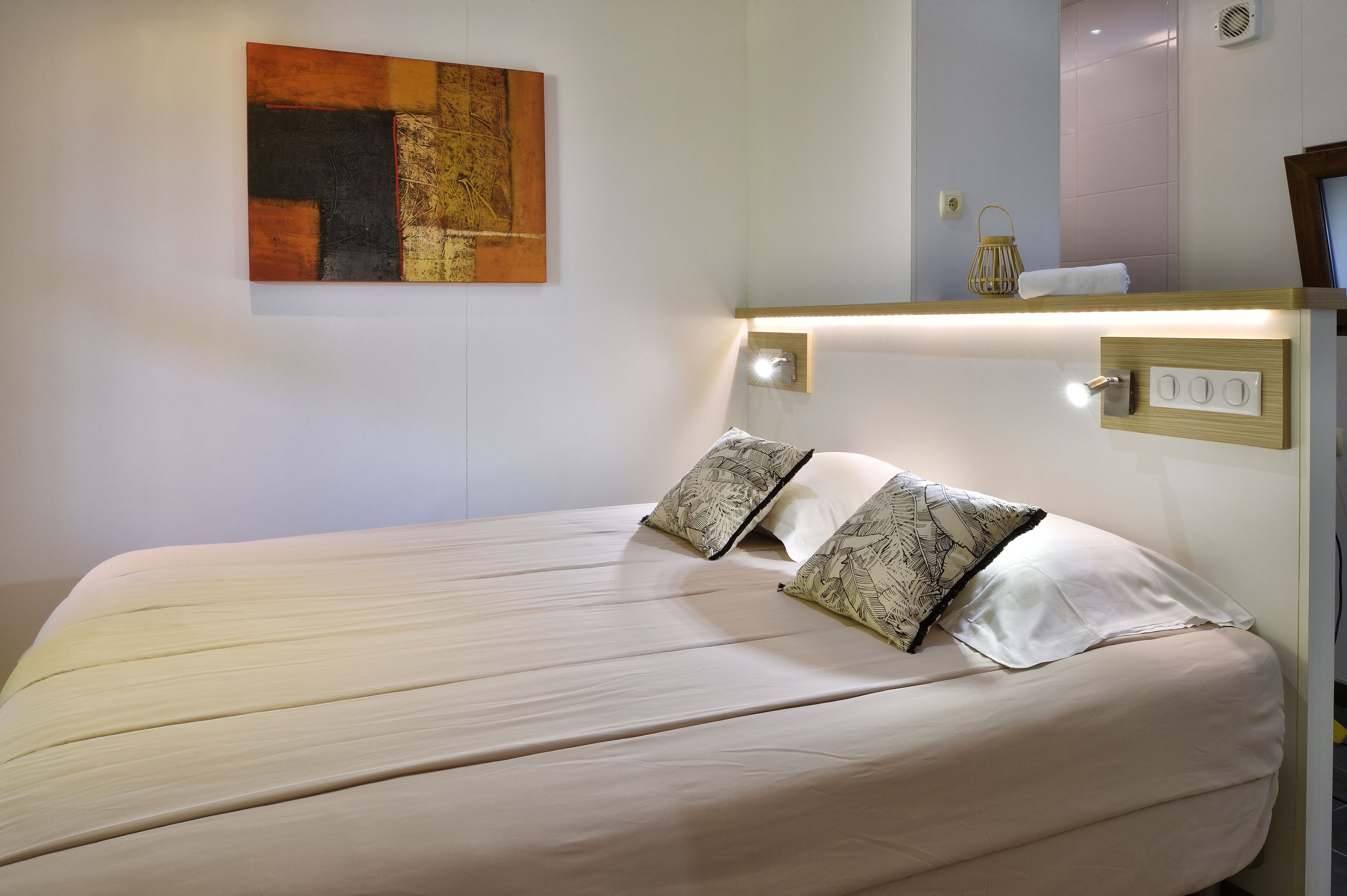 Un lit bien fait, recouvert d’une couvre-lit de couleur claire et agrémenté de deux coussins à motifs, un tableau abstrait accroché au mur et une table de chevet avec une lampe et un petit objet décoratif.