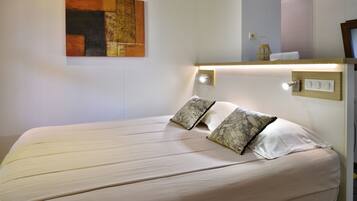 Un lit bien fait, recouvert d’une couvre-lit de couleur claire et agrémenté de deux coussins à motifs, un tableau abstrait accroché au mur et une table de chevet avec une lampe et un petit objet décoratif.