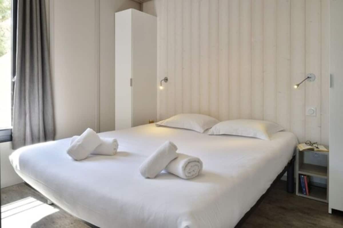Una cama bien hecha con sábanas blancas y toallas dobladas, una mesita de noche con un libro y gafas, y una ventana con cortinas.