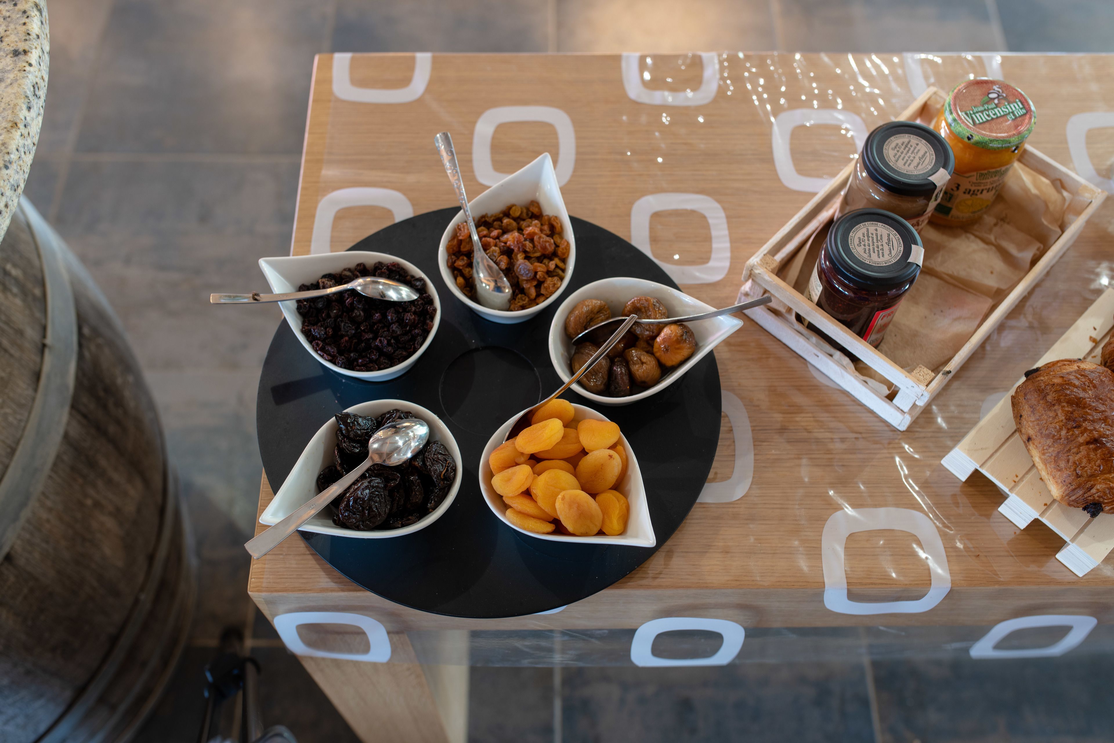 Una mesa con diversos alimentos y condimentos, incluyendo tazones de frutas secas y frutos secos, una bandeja con frascos y una rebanada de pan.