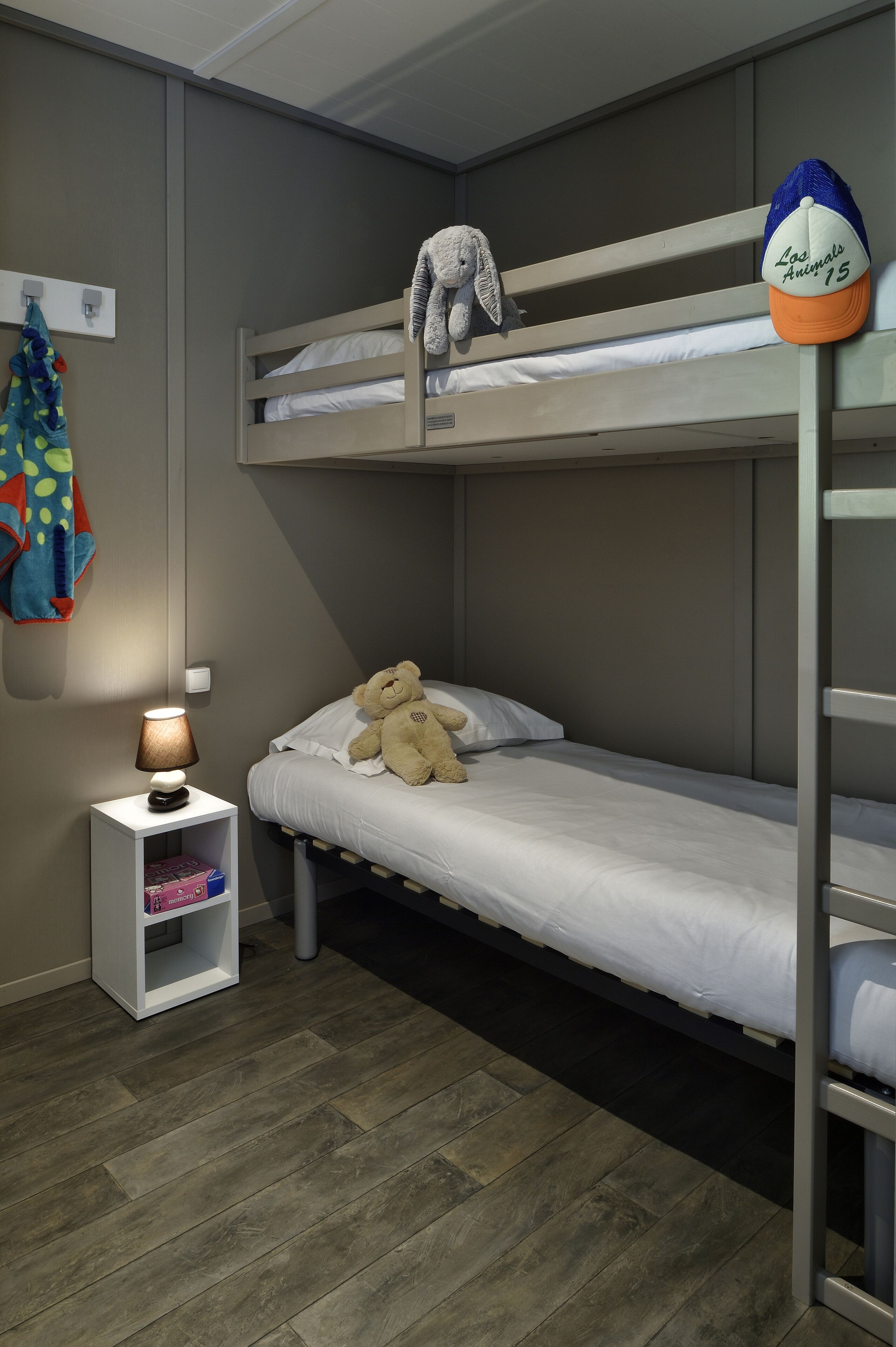 Une chambre avec un lit superposé, un ours en peluche sur le lit du bas, une lampe sur une table de chevet et une veste colorée accrochée au mur.