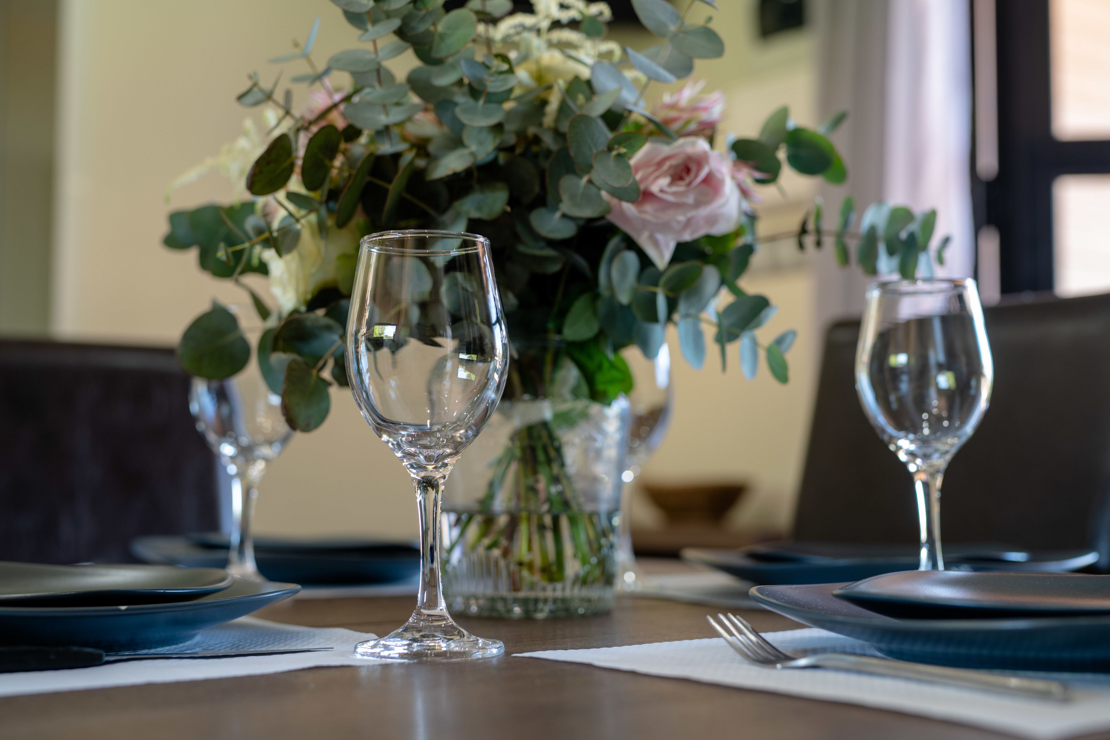 Een eettafel gedekt met blauwe borden, glazen wijnglazen en een vaas met bloemen.