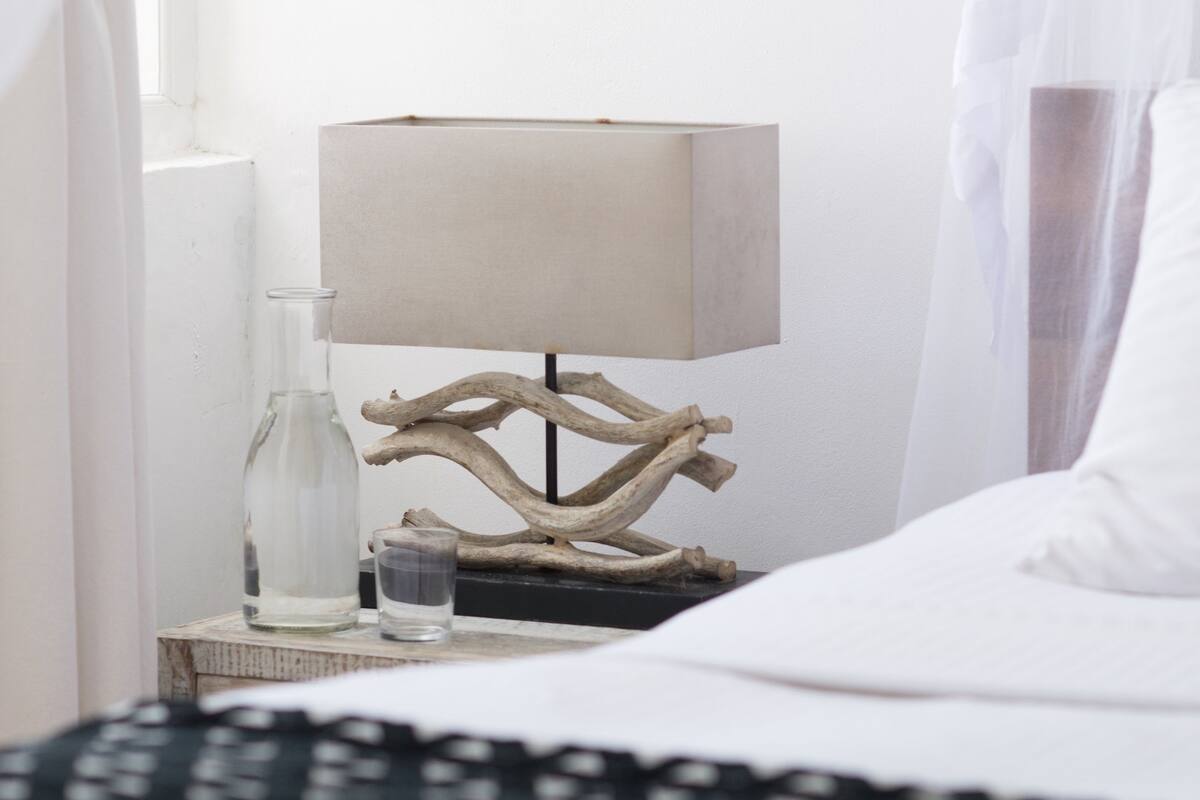 A neatly made bed with a white duvet, a bedside table with a lamp, a glass bottle, and a small glass.
