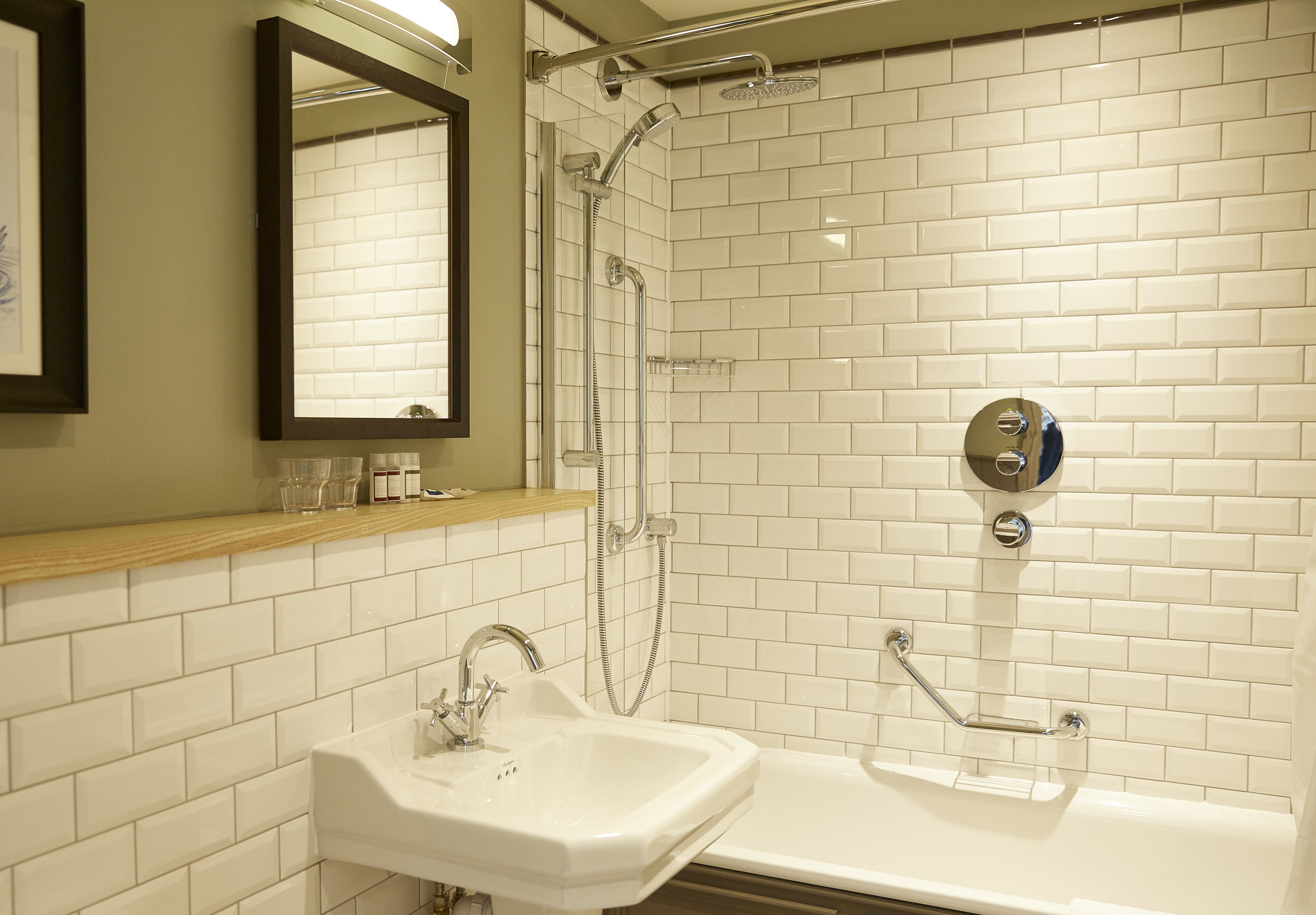 Un baño con azulejos blancos tipo metro, un lavamanos, un espejo y una ducha con cabezal de ducha manual.