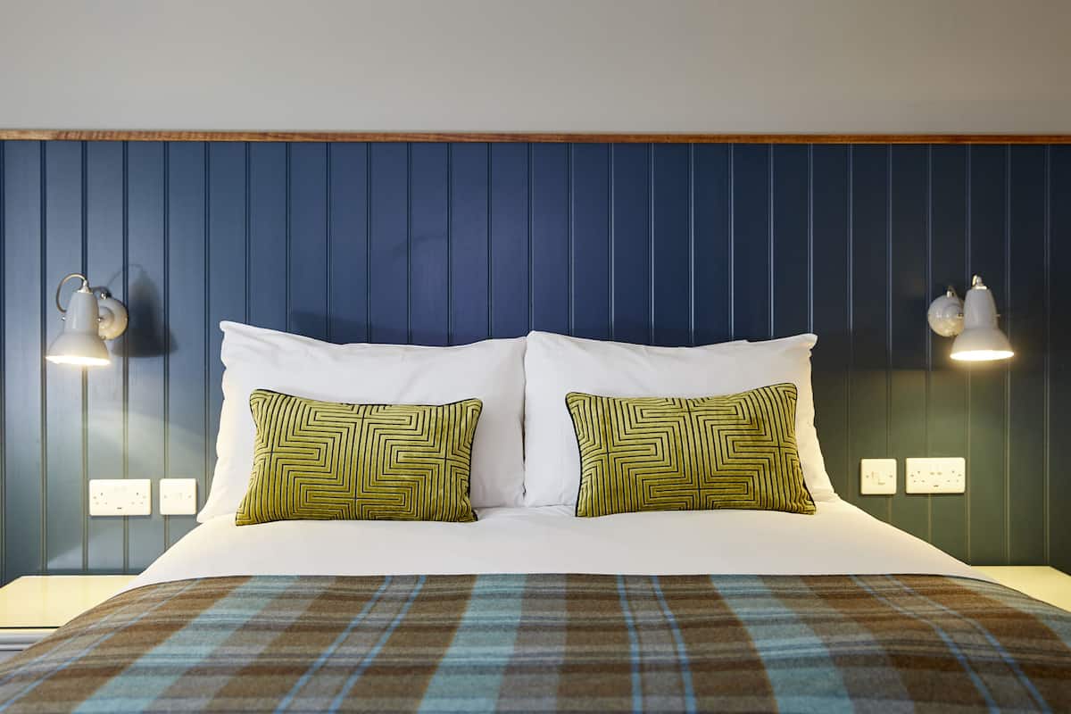 A hotel room with a large bed, two patterned pillows, a plaid bedspread, and wall-mounted lamps.