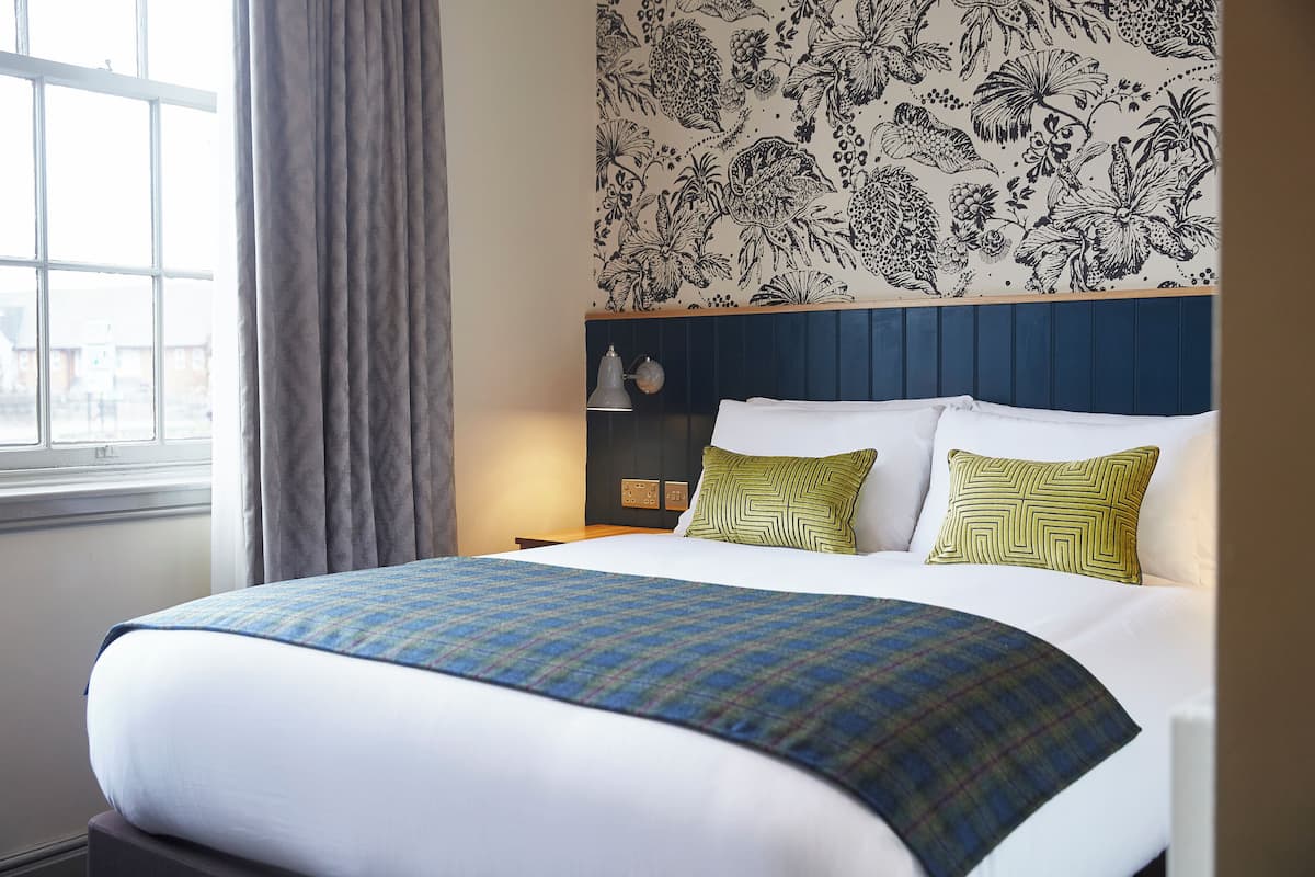 A neatly made bed with a plaid bedspread, two patterned pillows, and a floral wallpaper.