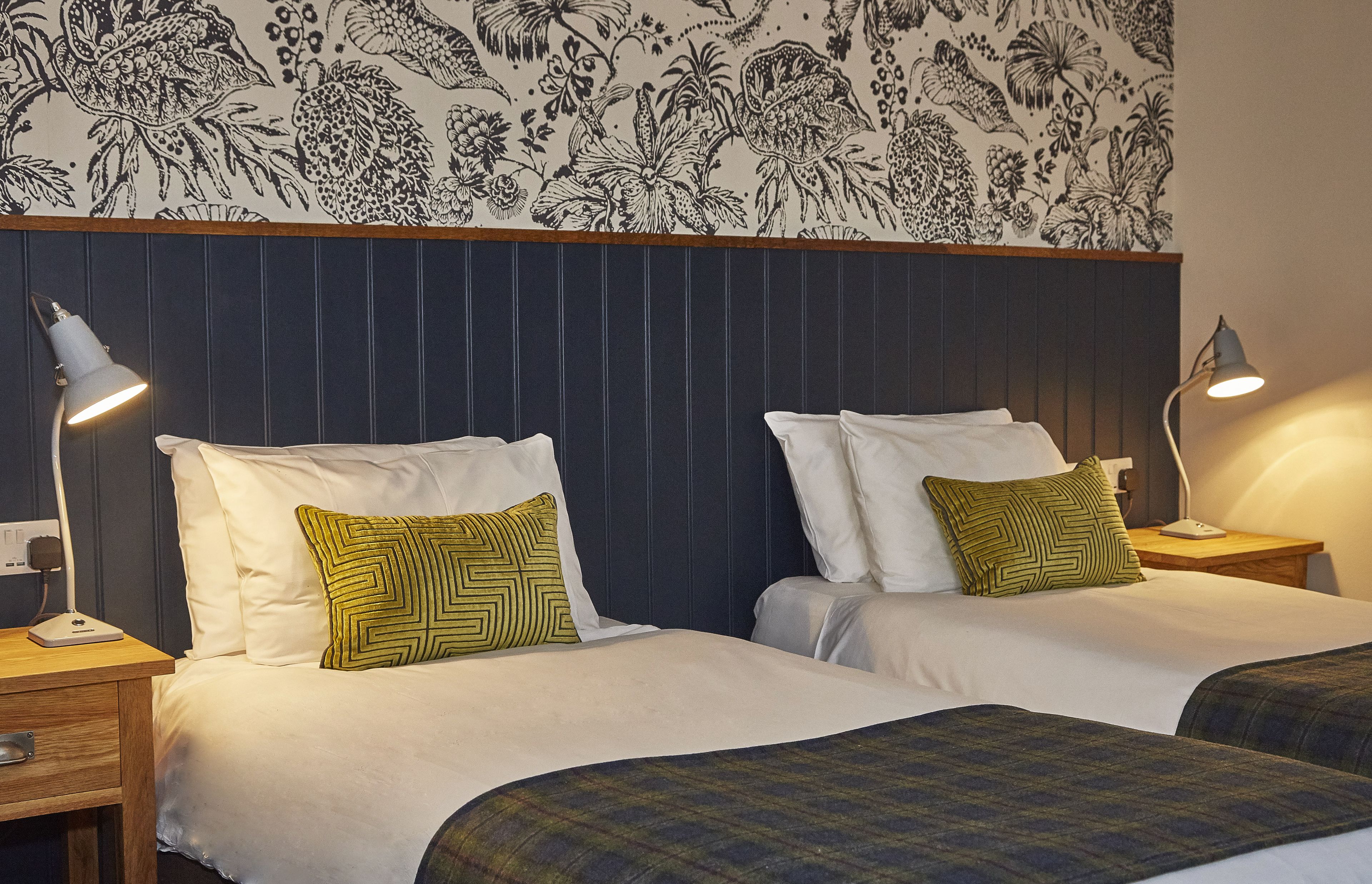 Habitación de hotel con dos camas, cada una con ropa blanca y almohadas estampadas en verde. Hay mesitas de noche con lámparas, y un papel tapiz con motivos florales sobre las camas.