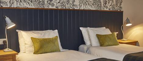 Une chambre d’hôtel avec deux lits, chacun doté de draps blancs et d’oreillers à motifs verts. On y trouve des tables de chevet avec des lampes, et un papier peint à motifs floraux au-dessus des lits.