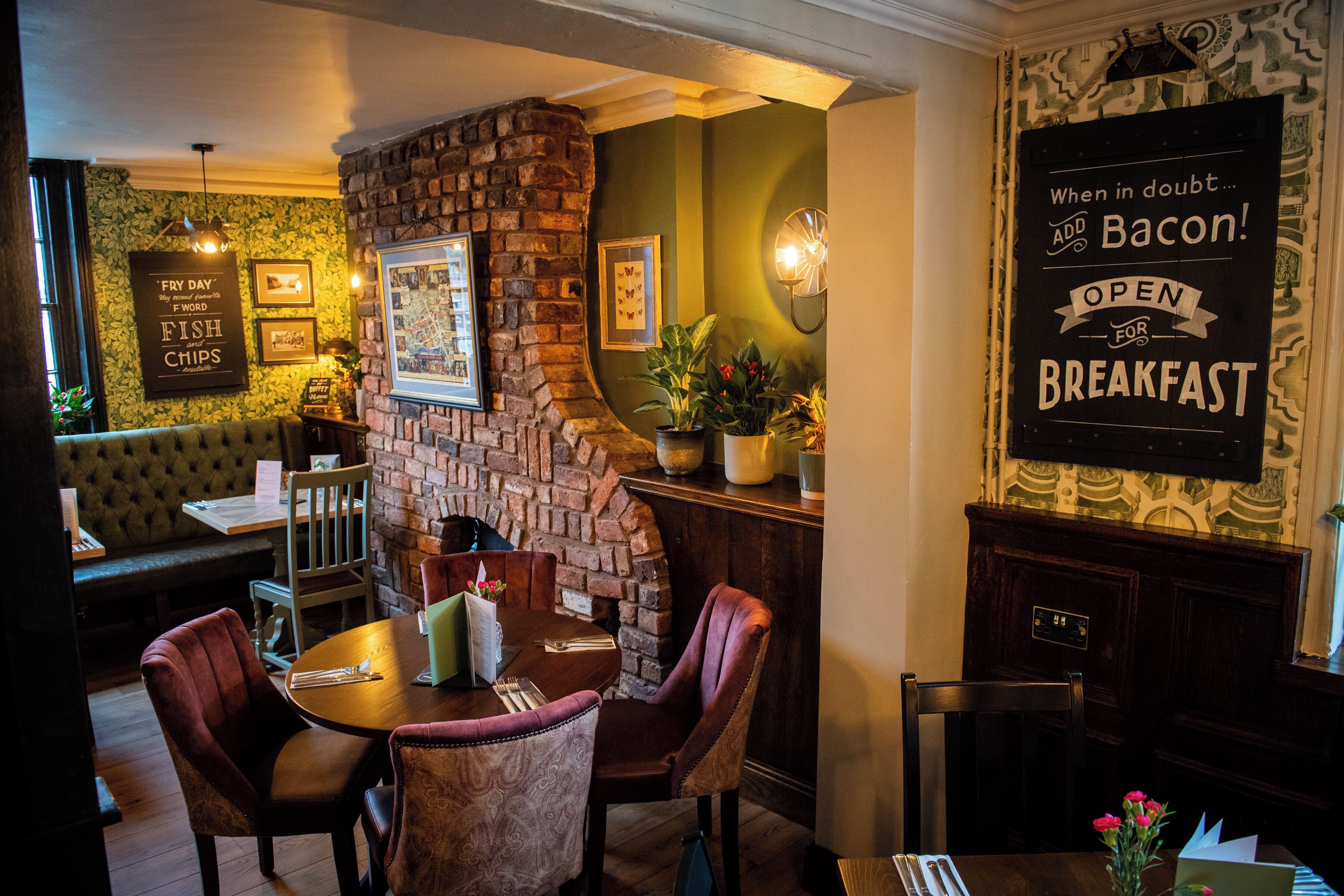 A restaurant with a brick wall, green walls, and a sign that says "When in doubt... add bacon! OPEN FOR BREAKFAST." There is a table with chairs, a bench, and a potted plant.