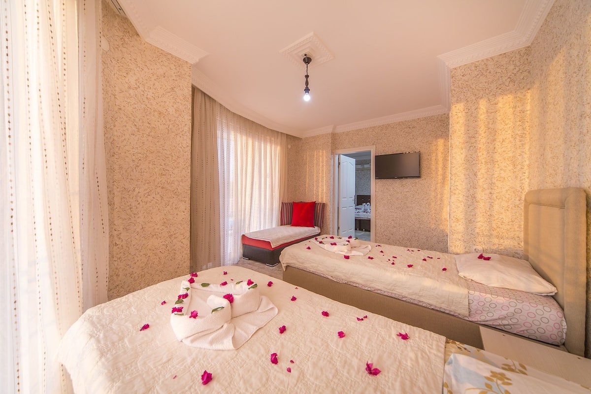 A hotel room with two beds, a sitting area with a red pillow, and a TV mounted on the wall.