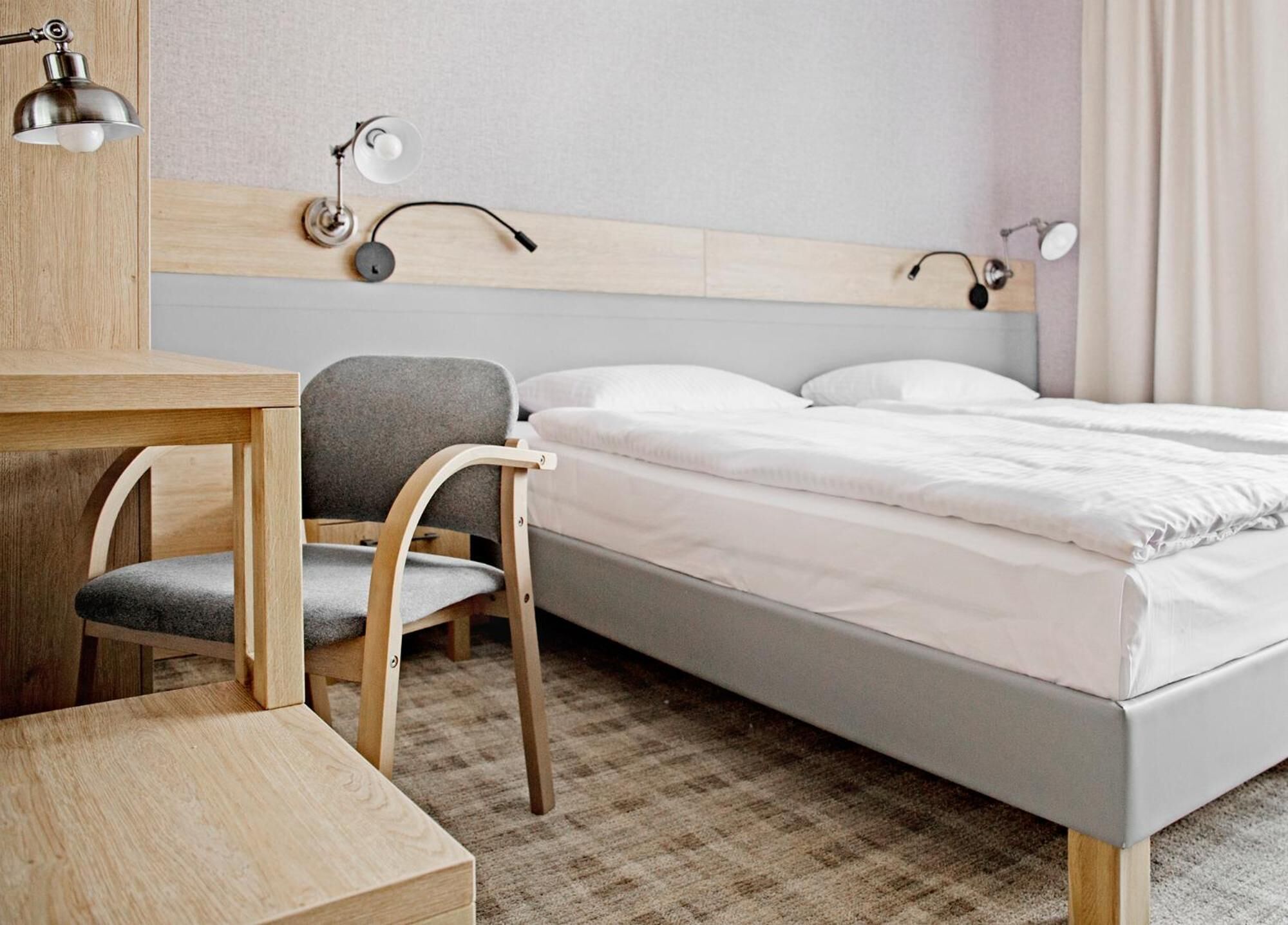 A hotel room with a wooden desk, a grey chair, a bed with white linens, and a headboard with wall-mounted lamps.
