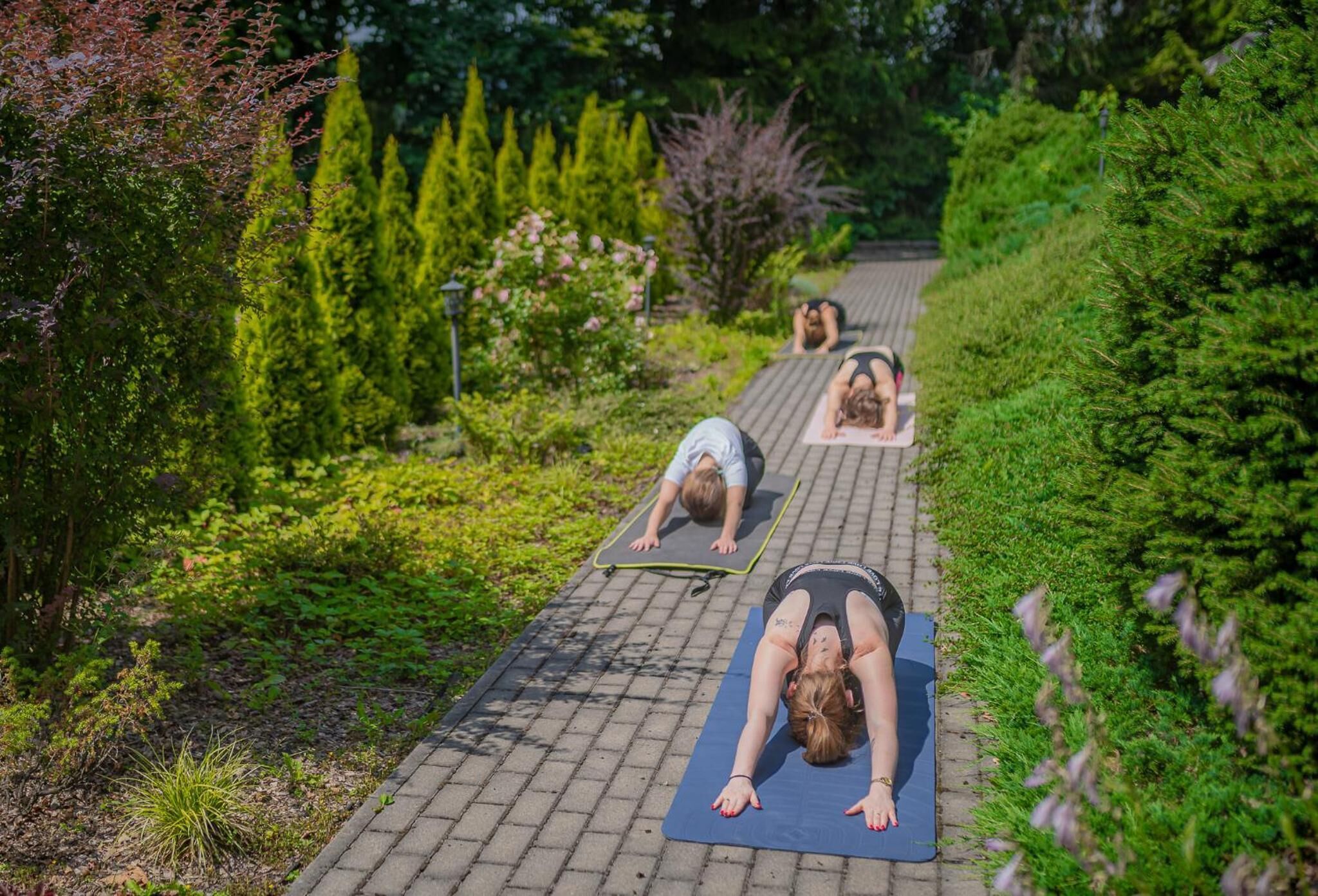 Drie personen doen yoga op matten in een open omgeving, met een verhard pad en groen.