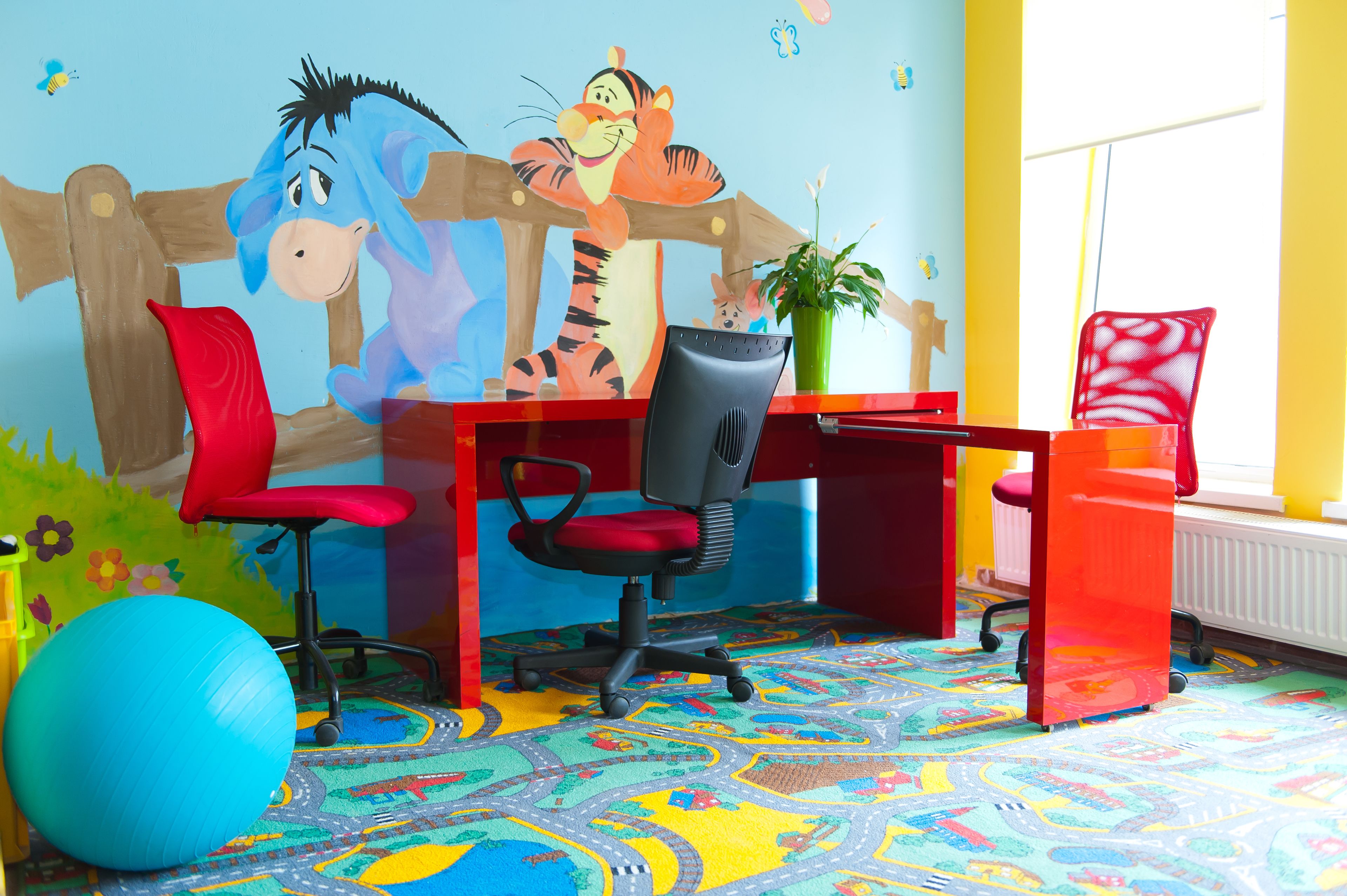 A child's playroom with a colorful carpet, a desk, two red chairs, a blue exercise ball, and a wall mural.