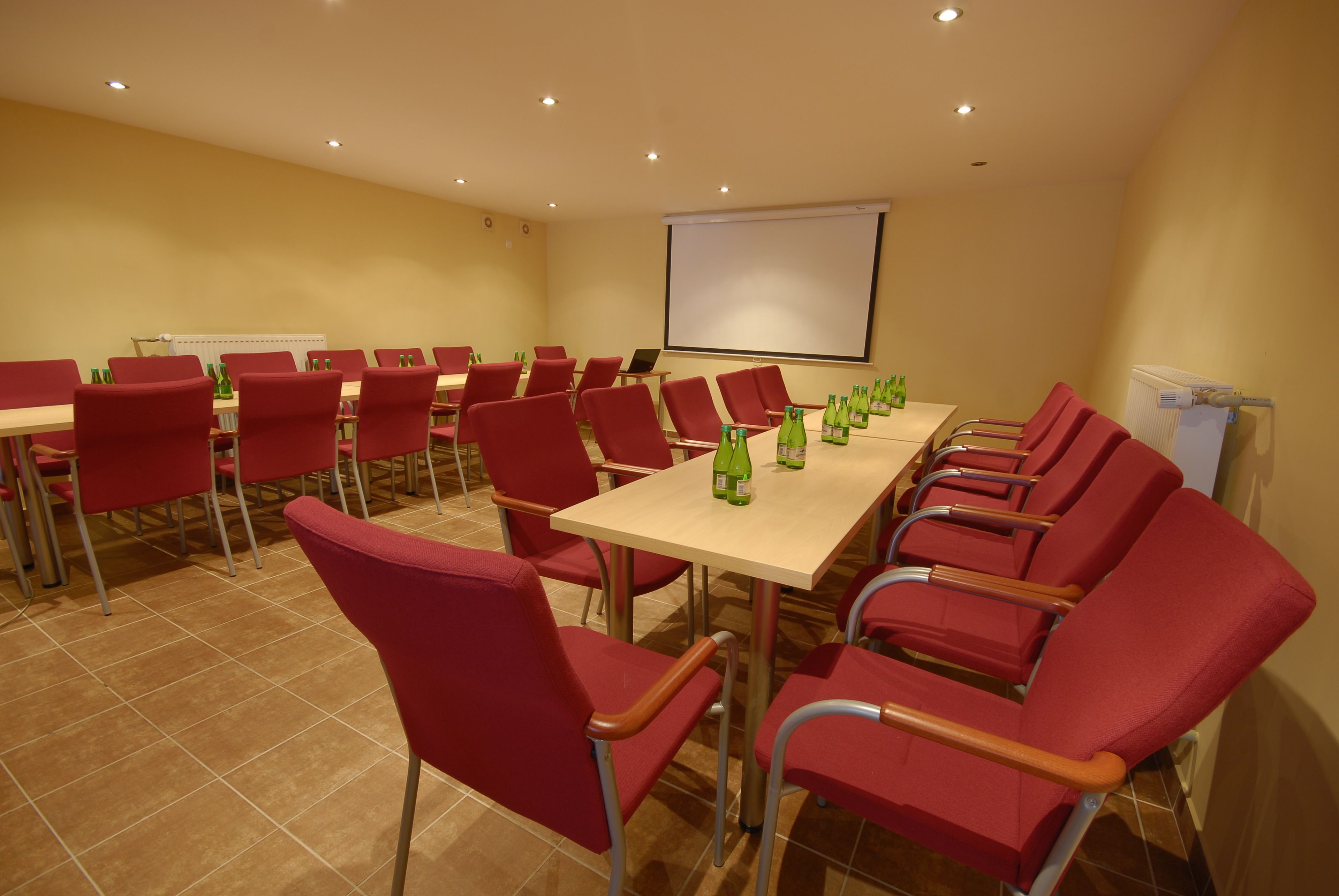 Un salón de conferencias con sillas y mesas rojas, una pantalla de proyector y botellas verdes sobre las mesas.