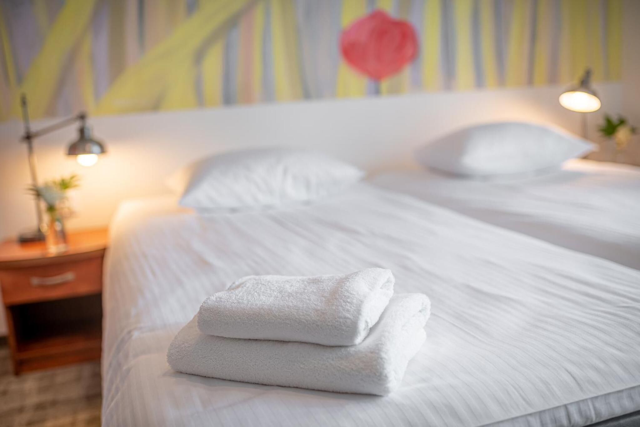 Una cama bien hecha con sábanas blancas y una toalla doblada, una lámpara de noche y una mesita de noche con una pequeña planta.