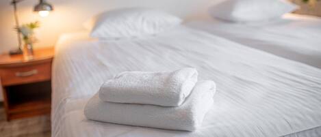 Una cama bien hecha con sábanas blancas y una toalla doblada, una lámpara de noche y una mesita de noche con una pequeña planta.