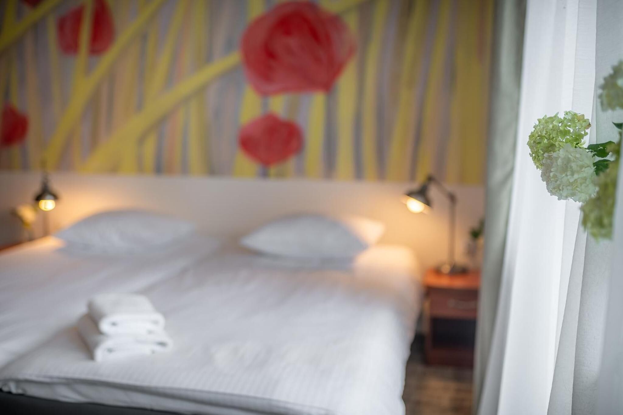 A neatly made bed with white linens and a folded towel, a bedside lamp, and a wall with a floral pattern.