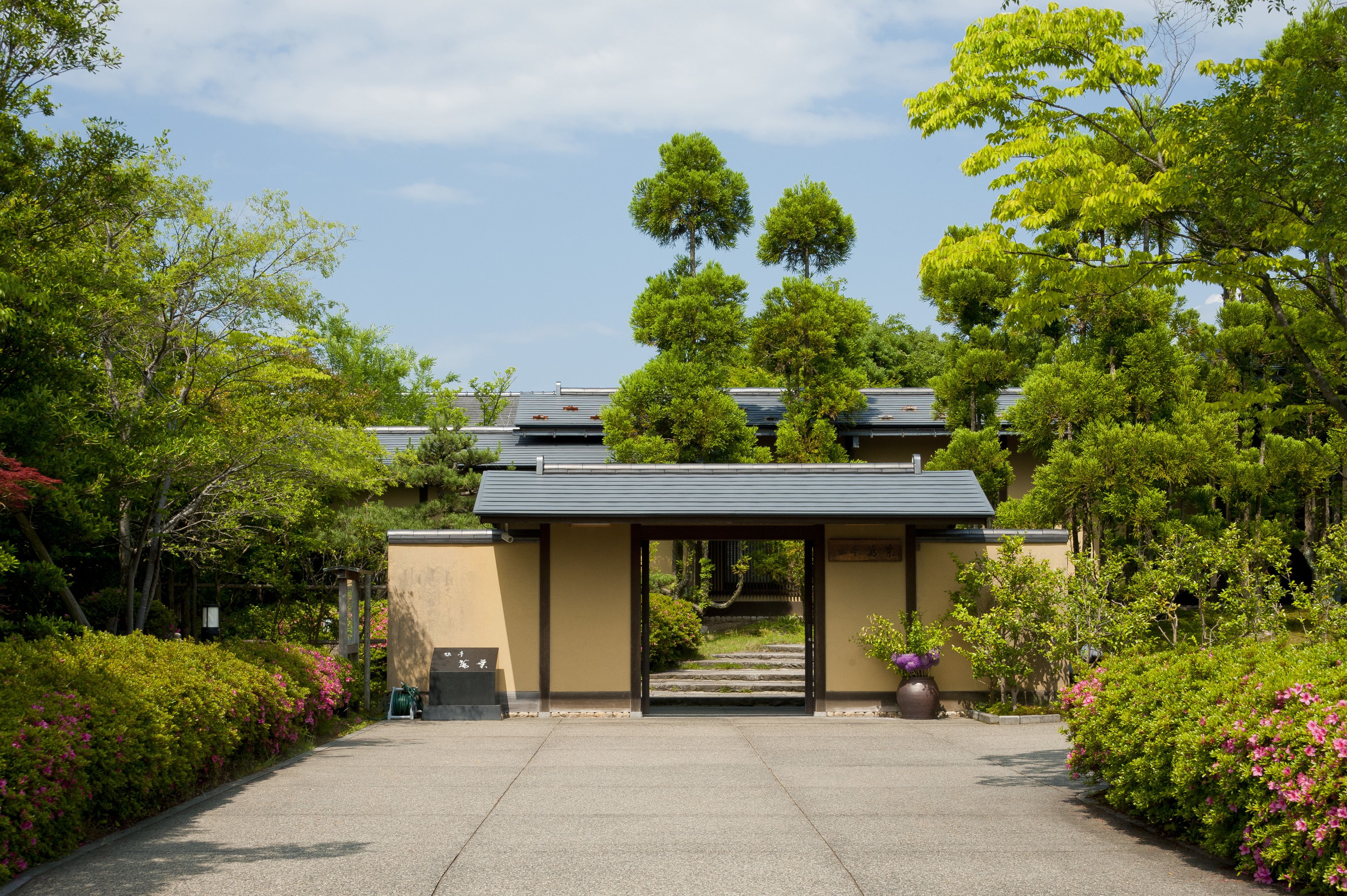A traditional Japanese building with a covered entrance, surrounded by lush greenery and flowering plants.