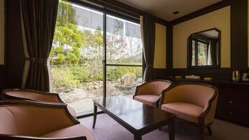 A hotel room with a large sliding glass door leading to a garden, two armchairs, a small table, and a dresser with a mirror.