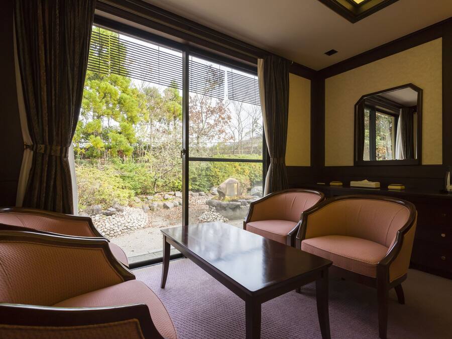 A hotel room with a large sliding glass door leading to a garden, two armchairs, a small table, and a dresser with a mirror.